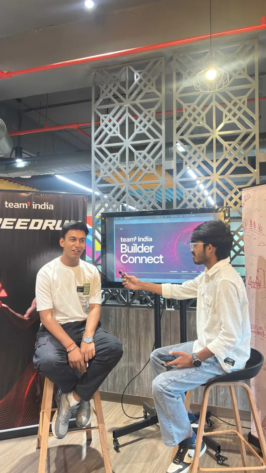 Two builders seated on stools recording an on-site conversation beside a presentation screen